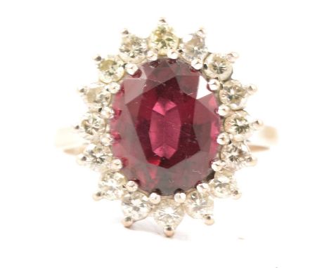 A garnet and diamond cluster ring, an oval mixed cut garnet 10.5mm x 8.5mm x 5.9mm, claw set and surrounded by sixteen brilli