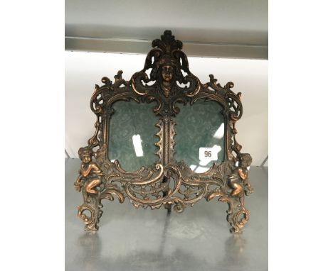 An early 20th century easel back copper plated metal double picture frame with mask finial and cherub mounts.