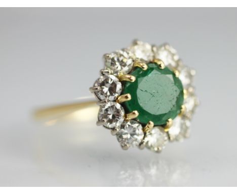 An emerald and diamond circular cluster ring, the central circular emerald, claw set in yellow gold and within a surround of 