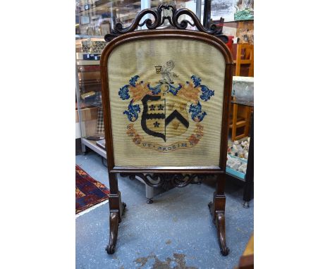 A Victorian rosewood firescreen with scrolling top above glazed panel set with a needlework coat of arms with motto 'Ut Prosi