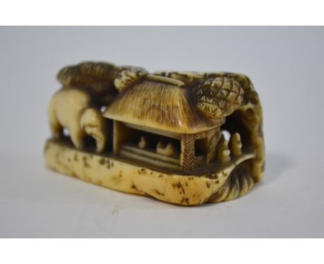 A stained ivory netsuke, carved as an elephant beside figures and two thatched dwellings, inscribed with two kanji on the bas