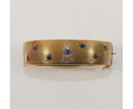 A 9 carat gold hollow hinged sapphire set bangle, with engine turned decoration, the seven round mixed-cut sapphires within g