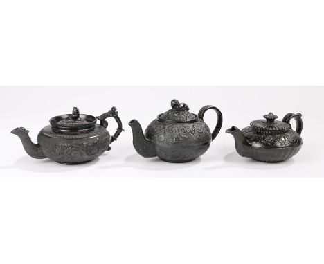 Three 19th Century black basalt bachelors teapots, the first surmounted by a reclining dog above a scroll and star burst body