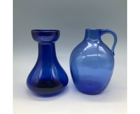 A pair of handblown blue glass pieces, including a jug and a vase. The jug features a classic round shape with a curved handl