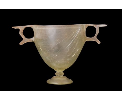 Ca. 200 BC. An elegant glass skyphos consisting of a deep cup supported by a pedestal foot, with two opposing handles adornin