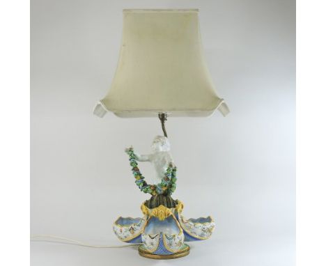 An Italian majolica figural table lamp and shade, modelled as a cherub holding a garland of flowers, above three scalloped bo