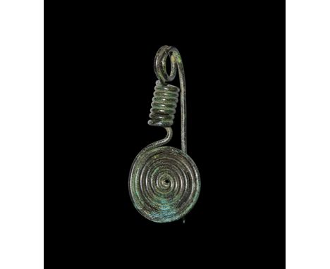Early 1st millennium BC.   A bronze fibula comprising a flat-coiled rod plaque with hooked catch to the underside, tubular-co