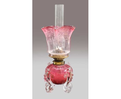 A Victorian cranberry glass oil lamp, the reservoir mounted on four fish shaped feet, with hinks duplex burner and cranberry 