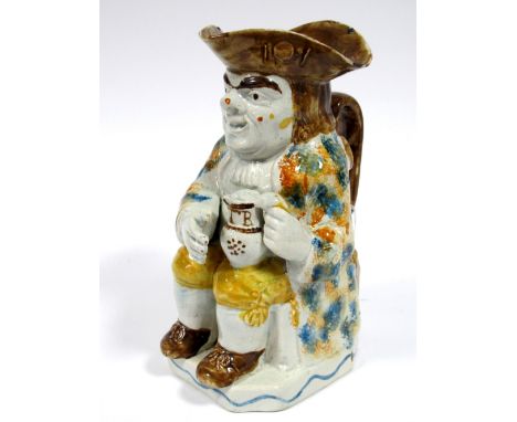 An Early XIX Century Prattware Toby Jug, the seated toper holding a jug of ale on his left knee, a pipe beside him, wearing a