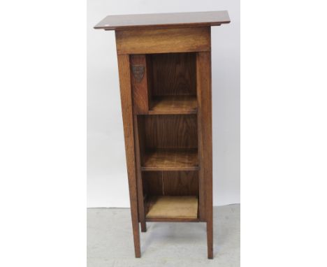 An Arts &amp; Crafts oak freestanding tapering bookcase with three shelves and small panel to one side with brass shield, eac