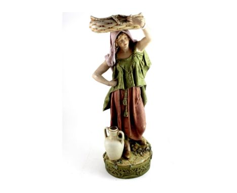 An early 20th century Royal Dux figure of a water carrier with water jar at her feet and basket on her head, supported on a s