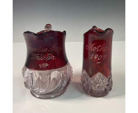 A pair of antique red and clear glass souvenir creamers, each steeped in historical charm. The first creamer, inscribed "Gran