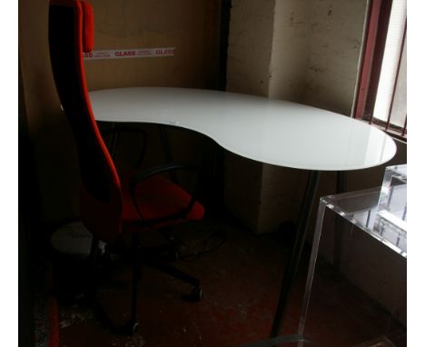 A modern desk with metal adjustable stand and kidney shaped opaque glass top;  an IKEA Markus swivel office chair, in orange 
