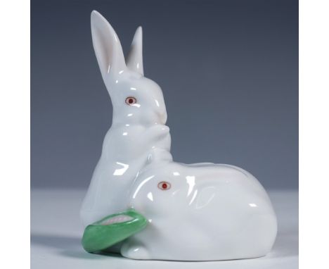 Herend porcelain figurine featuring two white rabbits, one seated upright and the other curled at its feet, sharing a green l