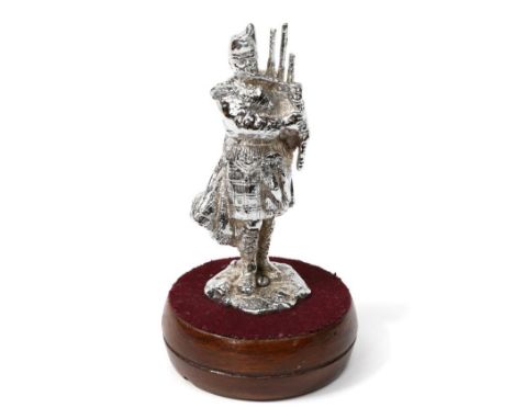 A Chrome on Brass Car Mascot modelled as a Scottish Piper, playing bagpipes and standing on a rocky base, mounted on a circul