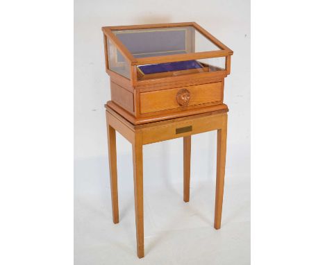 Light oak collectors display case on stand, the glazed top enclosing angled plinth, over single drawer with carved mask handl