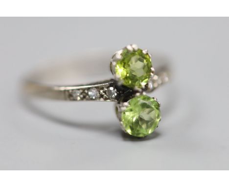 An 18ct and plat, two stone peridot crossover ring, with diamond set shoulders, size W, gross 2.7 grams.CONDITION: Some wear 