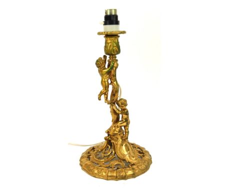 An early 20thC gilt metal table lamp, in the rococo style, with two cherubs climbing tree, on a circular foot, with cream sha