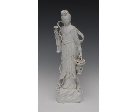 A Chinese Blanc de Chine figure, of Guanyin, she stands in flowing robes and sash, holding a basket of flowers in one hand an