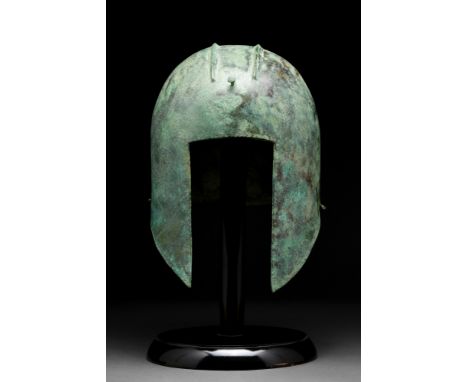 Ca. 600 BC.A finelly modelled helmet made of hammered bronze sheet, with a rectilinear opening for the face, long, pointed ch