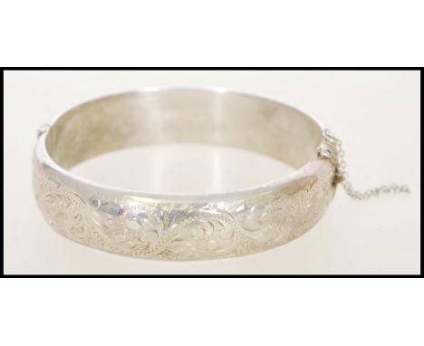 A 1980's silver bangle bracelet with a hinge opening, safety chain and floral engraving. Weight approx 23g. Measures 7 cm dia