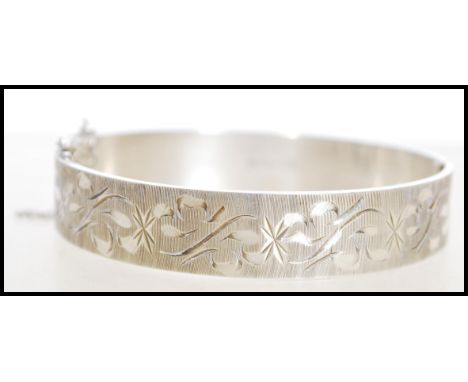 A 20th century hallmarked silver bangle bracelet with floral engraving and a hinge opening and safety chain. Hallmarked Birmi