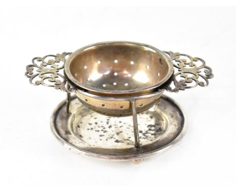 A George V hallmarked silver tea strainer on stand, circular with pierced foliate handles, the stand on three ball feet, Harr