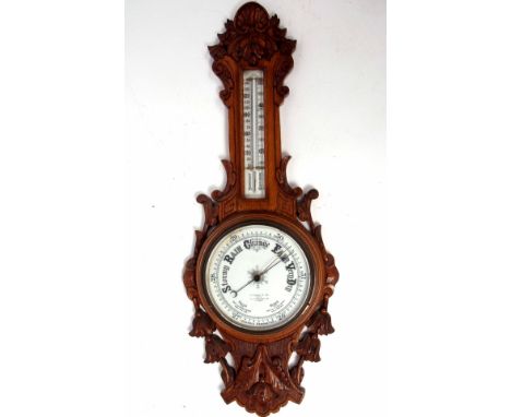 Late 19th century carved oak aneroid wheel barometer with carved and pierced foliate detail to a twin scale porcelain backed 