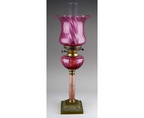 An early 20th Century oil lamp with ruby glass wyvern twisted shade and reservoir, on mottled marble column with brass raised