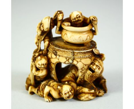 A JAPANESE CARVED IVORY NETSUKE, depicting a man crouching beneath a table with a cauldron on the surface, surrounded by a gr