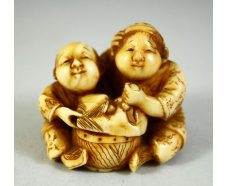 A JAPANESE CARVED IVORY NETSUKE, depicting two seated children holding a noh mask over a drum, 4cm wide.