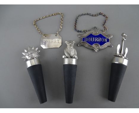Three white metal bottle stoppers in the form of a seated teddy bear, an anchor and the sun, a white metal and blue enamel bo