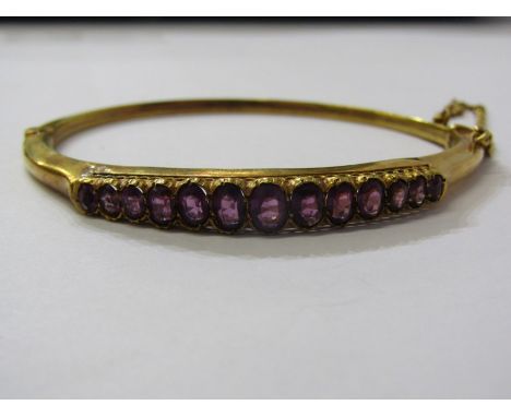 YELLOW METAL TESTS GOLD, HINGED AMETHYST BANGLE, line of double graduated oval cut amethysts in heavy yellow metal (testing g