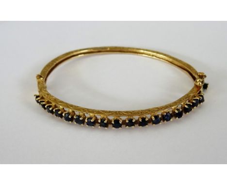 A gold and sapphire set oval hinged bangle, the front claw set with a row of circular cut sapphires, otherwise with scrolled 