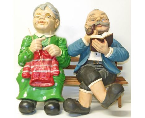 Painted plaster figure group of a lady and gentleman sitting on a bench knitting and smoking a pipe.