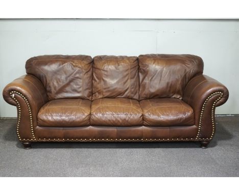 A leather four seater sofa with brass studding to arms and apron, upon turned wooden feet, 94cm high x 245cm wide x 102cm dee