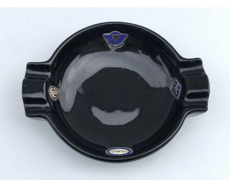 A black glazed pottery ashtray bearing four inset part enamel badges for Morris, MG, Wolseley and Riley, the ashtray with sha