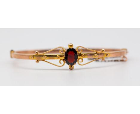 An early 20th century garnet set 9ct gold bangle, comprising a  central oval mixed cut garnet within scrolled shoulders, widt