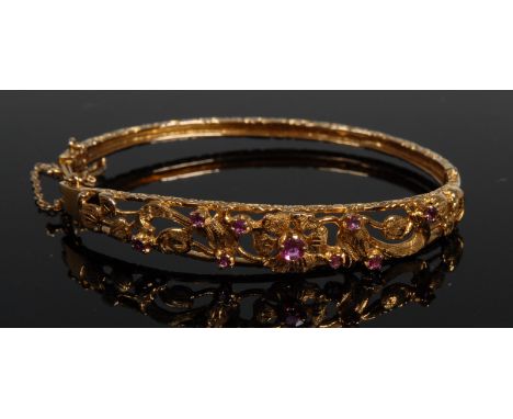An Art Nouveau open cast hinge bangle, central flower head set with a single round pink ruby, flanked by open scrolling sinuo