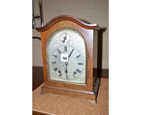 An Edwardian inlaid mahogany mantel clock, the serpentine top above arched panel dial with roman numerals, raised on brass br