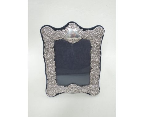 A silver photograph frame having extensive scroll repousse decoration and a plain cartouche with navy velvet surround and rev