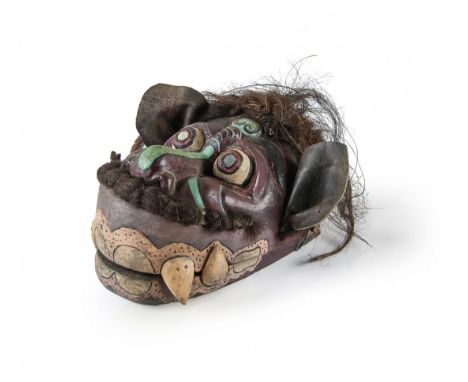 A face mask in wooden fabrication with dark brown  and light green coloration. The mask has black  hair on top of the head an