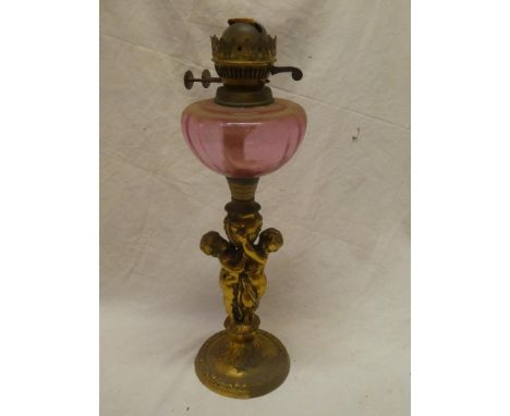 An unusual brass oil lamp, the stem modelled as two cherubs below a cranberry-tinted glass font
