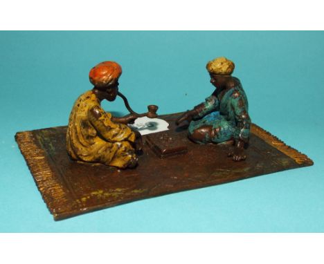 A painted metal group, of two eastern figures playing chess, on a rug, 11 cm in length