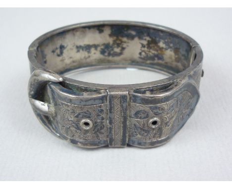A Victorian white metal hinged bangle, modelled in the form of a buckled belt, with foliate engraved face, 27.4g