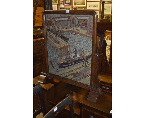 An early 20th century carved oak fire screen set with woolwork tapestry depicting a harbour scene with ships and industrial b