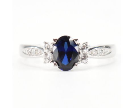 A hallmarked 9ct white gold synthetic sapphire and CZ ring. The ring set with an oval cut synthetic sapphire flanked by illus