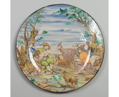 A large Italian Maiolica charger. Decorated in colours with a rural landscape scene with a goat herder playing bagpipes and w