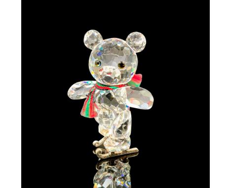 A clear crystal figure of a bear wearing a red and green scarf made of fabric that is on ice skates with melon-colored eyes. 