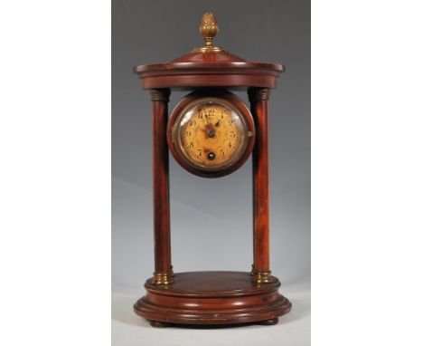 An early 20th Century mahogany portico mantel clock raised on a round plinth base, the round clock face raised between four c